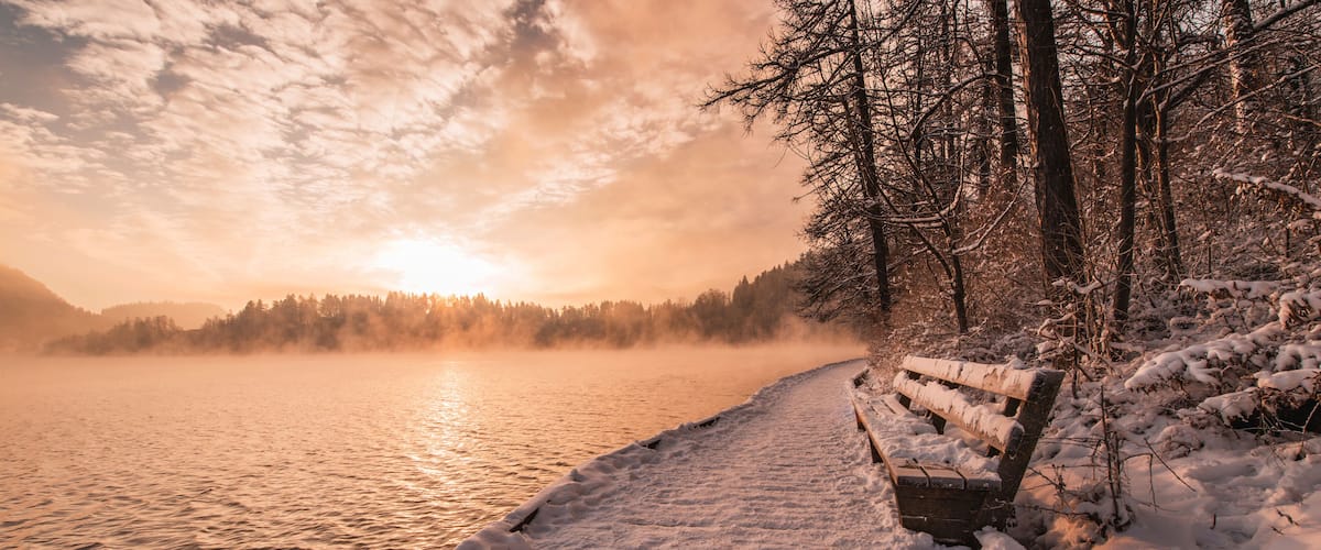 A snow-covered bench by a lake at sunset.