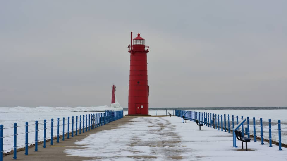 A red lighthouse on a snowy pier with blue railings and benches.