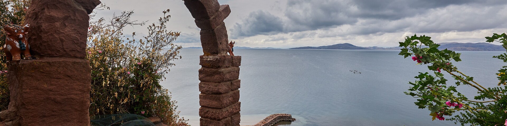 Un arco de piedra adornado con caballos, un banco de madera y vistas a un lago tranquilo con montañas al fondo.