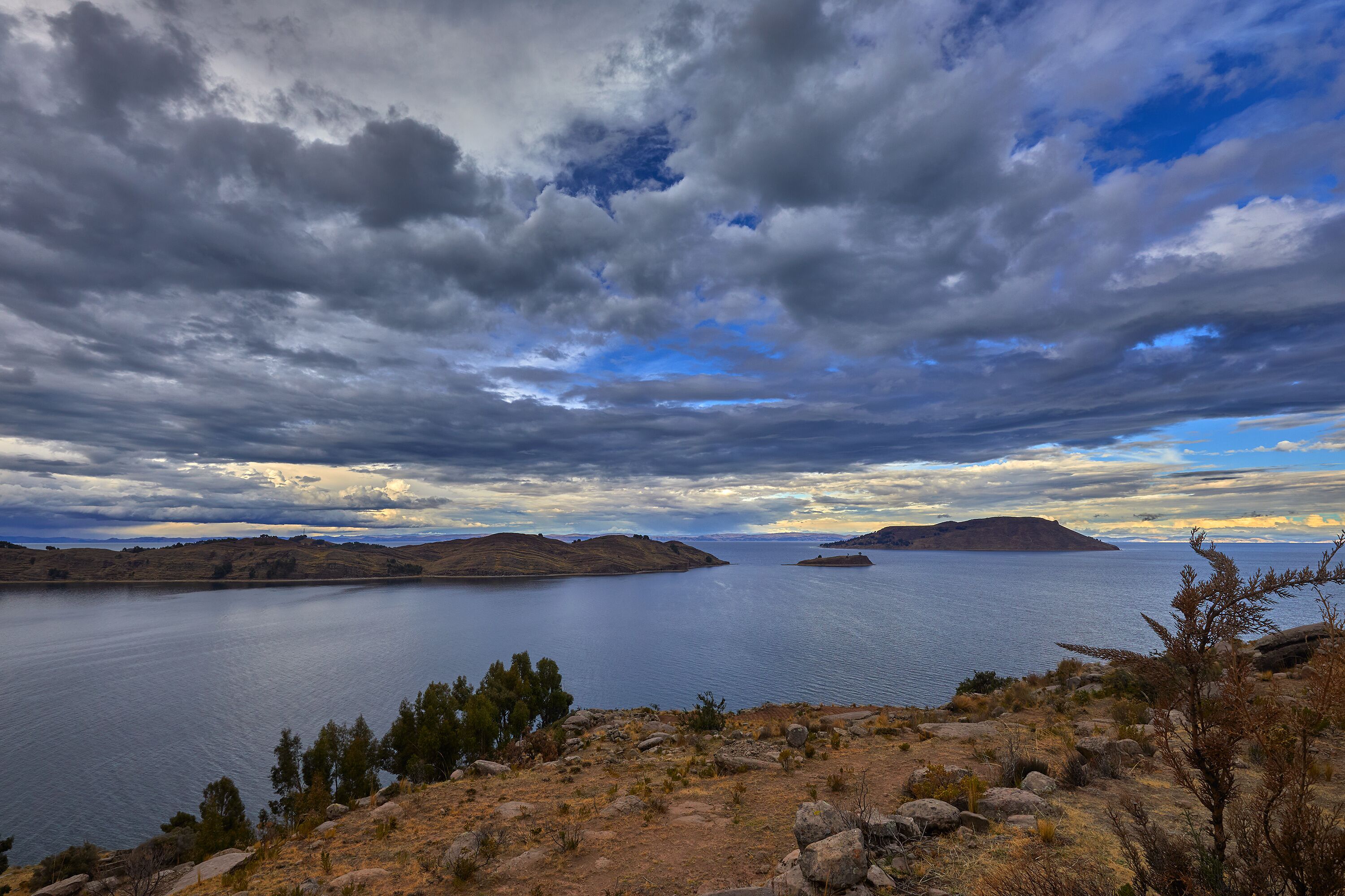 A scenic view of Lake Titicaca’s northern side from the Capachica Peninsula, showing Andean hills, deep blue waters, and traditional farmland under dramatic stormy skies.
