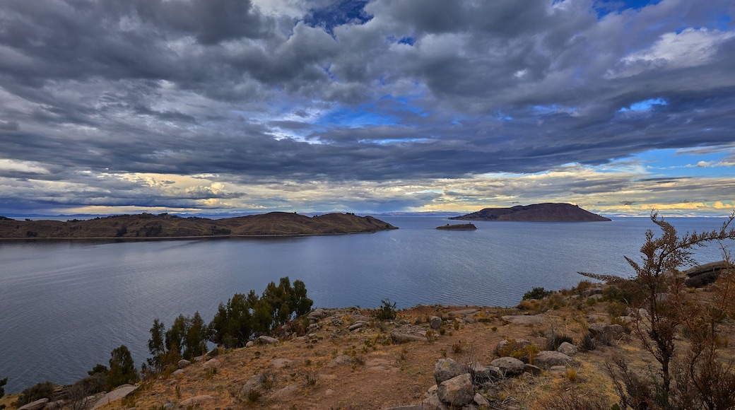 A scenic view of Lake Titicaca’s northern side from the Capachica Peninsula, showing Andean hills, deep blue waters, and traditional farmland under dramatic stormy skies.