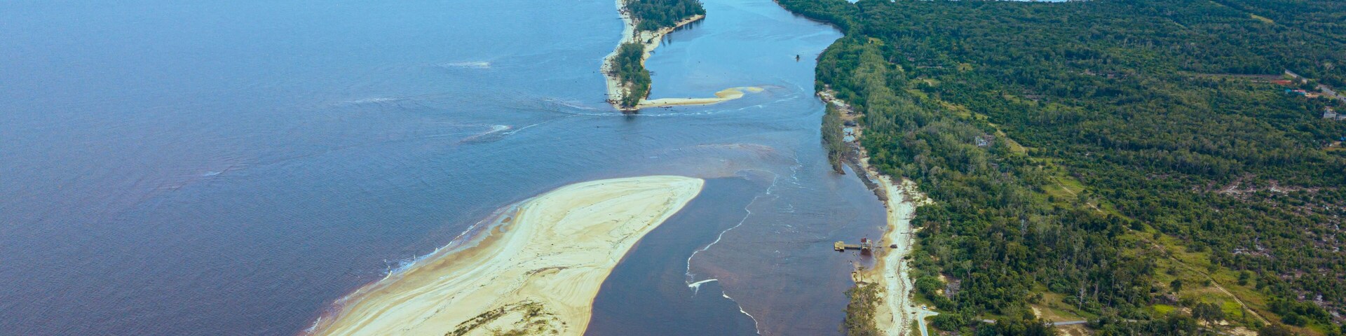 Vista aérea de una zona costera con una extensa playa arenosa, un río o bahía, y una región boscosa con algunas edificaciones.