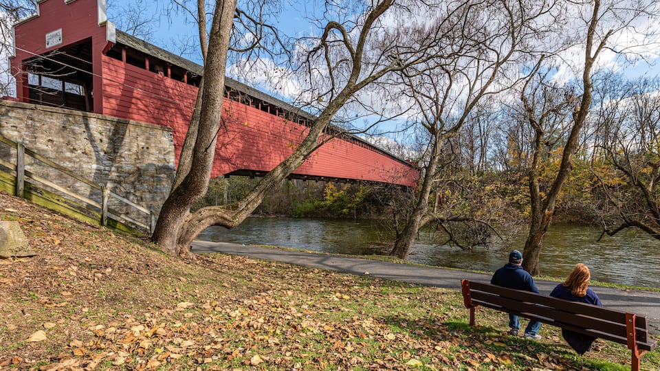 A red covered bridge, a stone wall, a river, and a park bench with two people sitting.