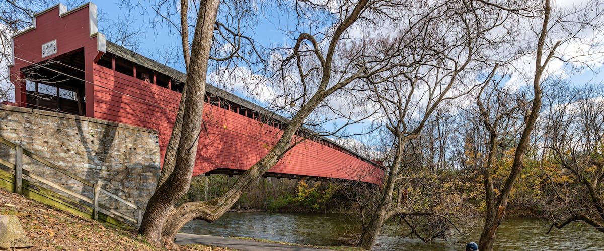 A red covered bridge, a stone wall, a river, and a park bench with two people sitting.
