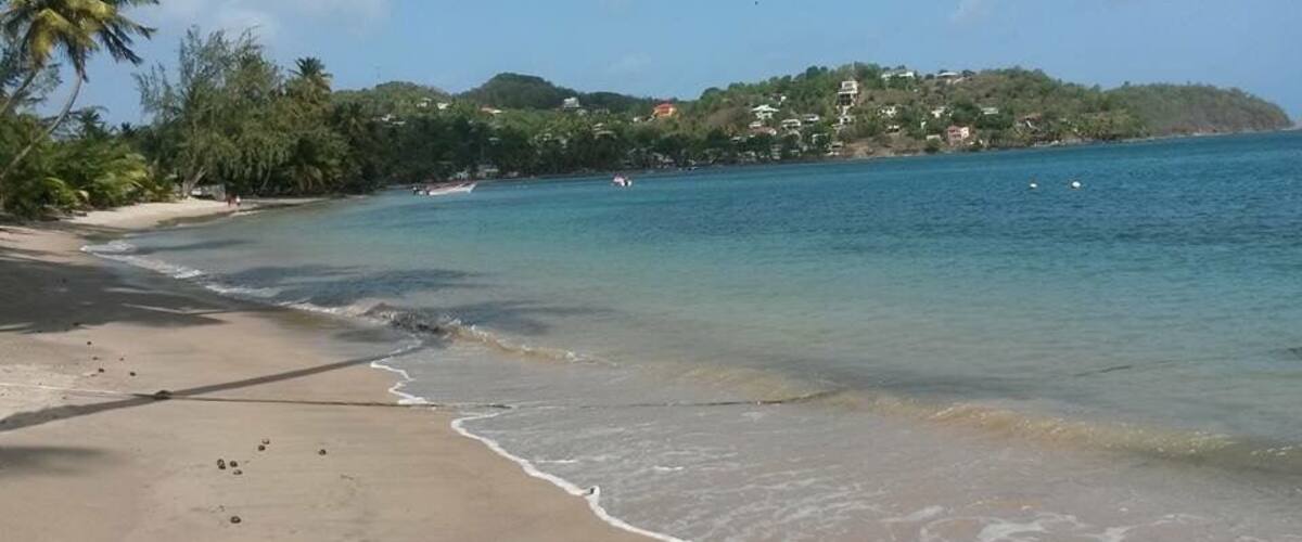 A beach with clear turquoise water, sandy shore, and a hill with buildings in the background.