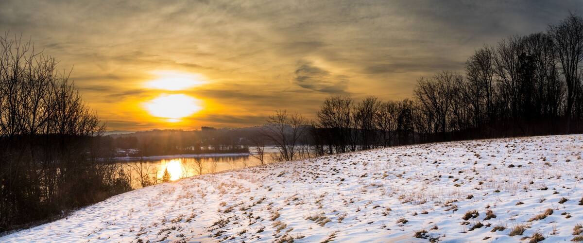 A snow-covered landscape with a sunset over a body of water and trees.
