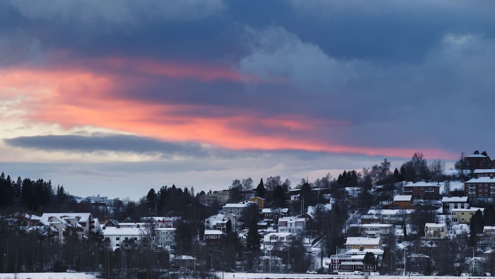 A snowy residential area with houses and trees under a colorful sunset sky.