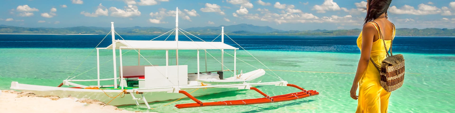 Une femme vêtue d’une robe jaune se tient sur une plage de sable, dominant une mer turquoise et limpide, avec un bateau traditionnel ancré à proximité.