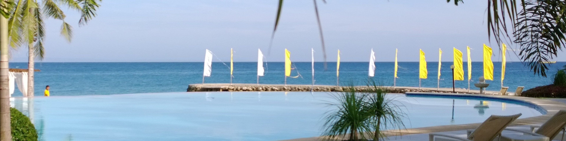 A pool with lounge chairs, palm trees, and flags by the ocean.
