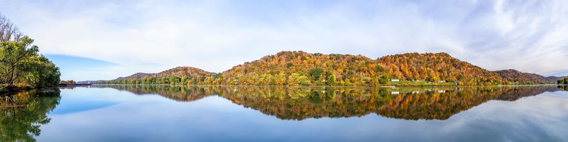 Hills covered in colorful fall foliage are reflected on the still water of the beautiful Ohio River. Monroe County, Ohio is viewed from the river bank at Paden City, West Virginia.