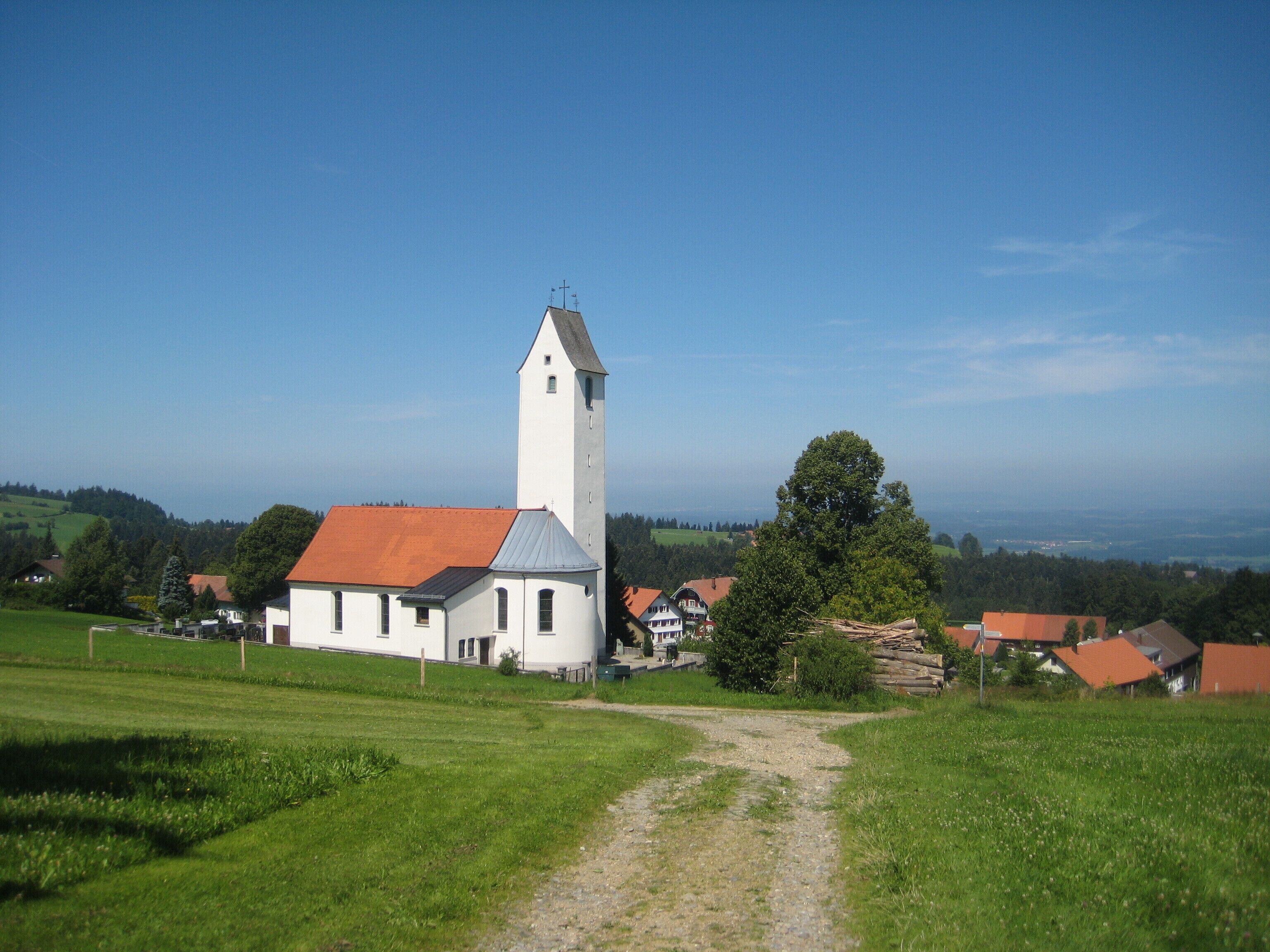 A white church with a tall tower and red roof, surrounded by green fields and houses.