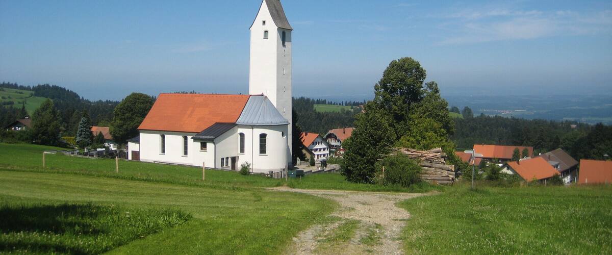 A white church with a tall tower and red roof, surrounded by green fields and houses.