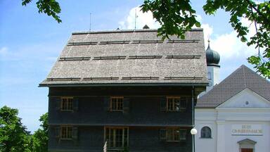 A large, dark-colored building with a steeply pitched roof and a smaller adjacent building with a white facade and a dome.