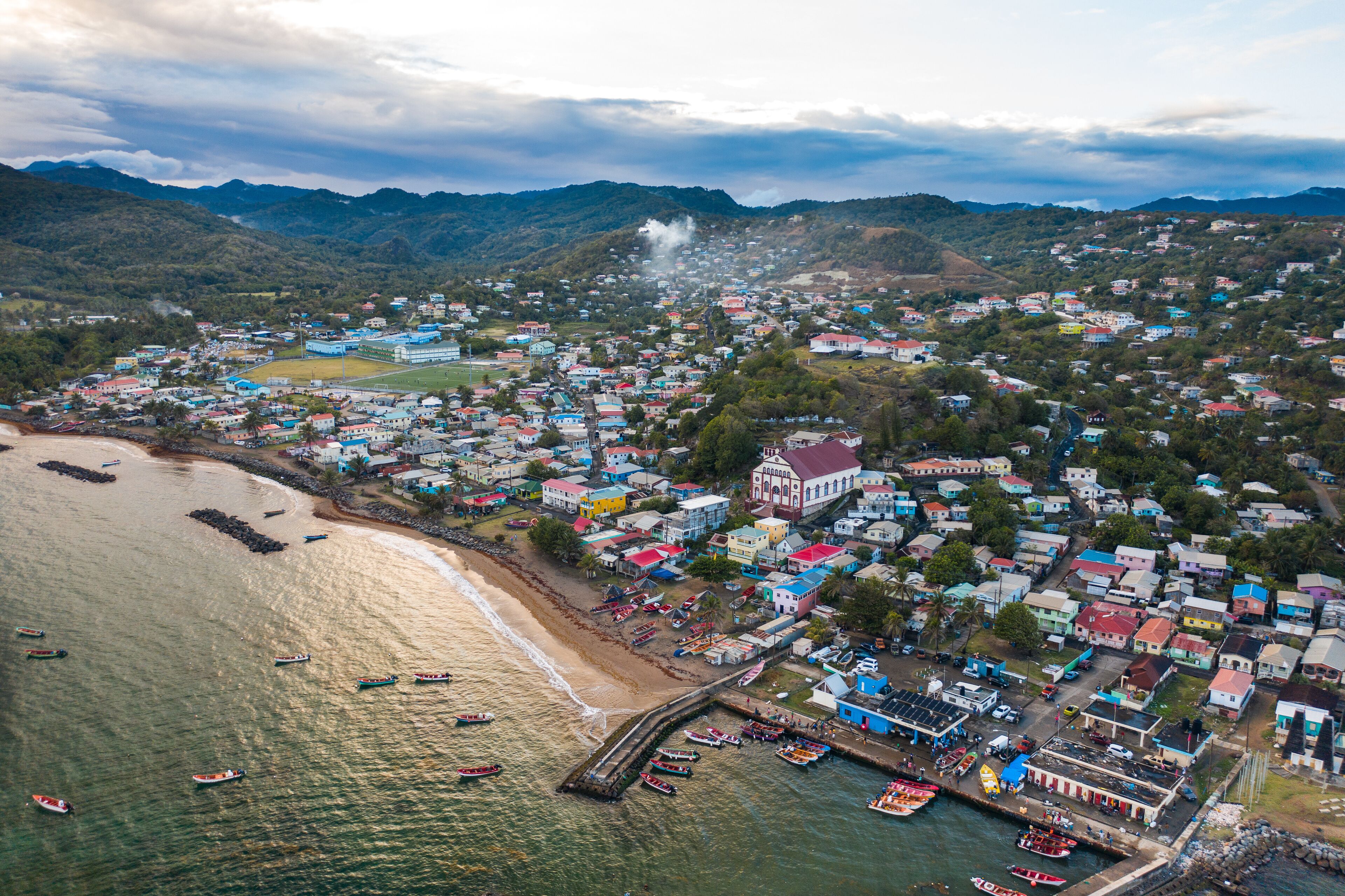An aerial view of a coastal town with colorful buildings, a beach, boats, and mountains in the background.