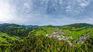 A mountainous landscape with a village nestled in a valley, surrounded by forests and green fields.