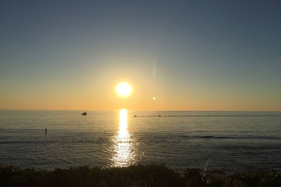 A sunset over a calm sea with a boat and a person standing on the shore.