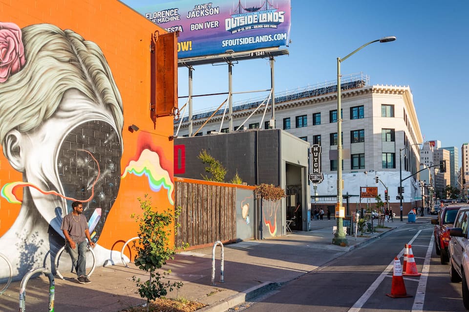 A mural of a woman's face on a building, a street with cars, and a billboard advertising an event.