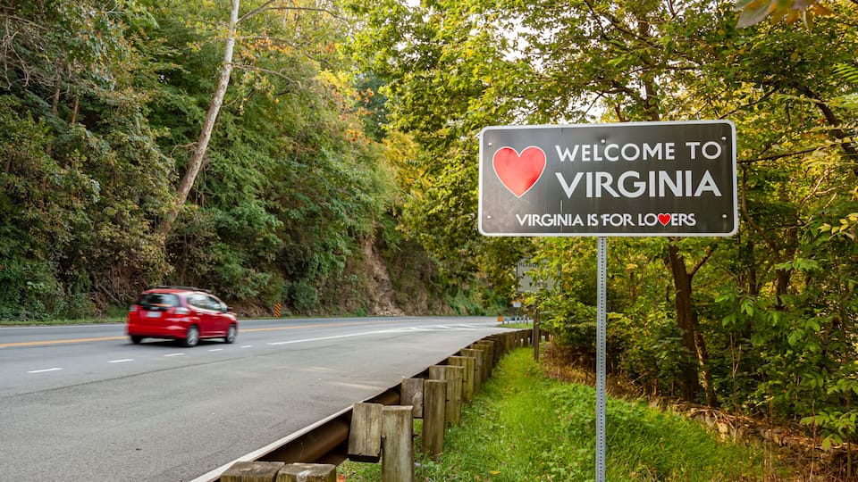 A road sign with 'Welcome to Virginia' and 'Virginia is for Lovers' text, surrounded by trees and a red car on the road.