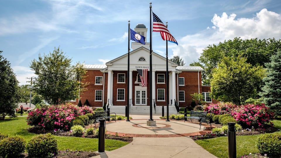 A town hall with a brick facade, two flags, and a landscaped garden with benches.