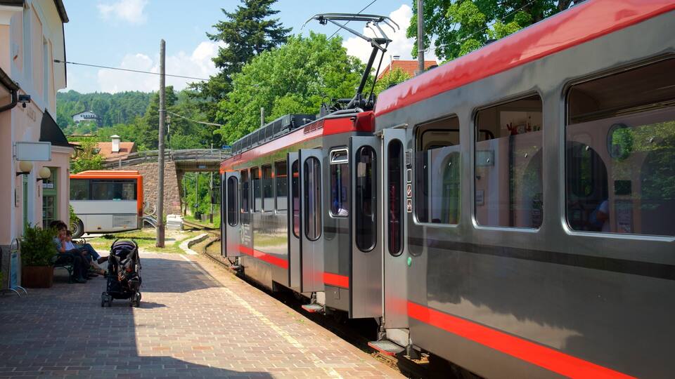 A train station with a red and silver train, a bus, and a pedestrian walkway.