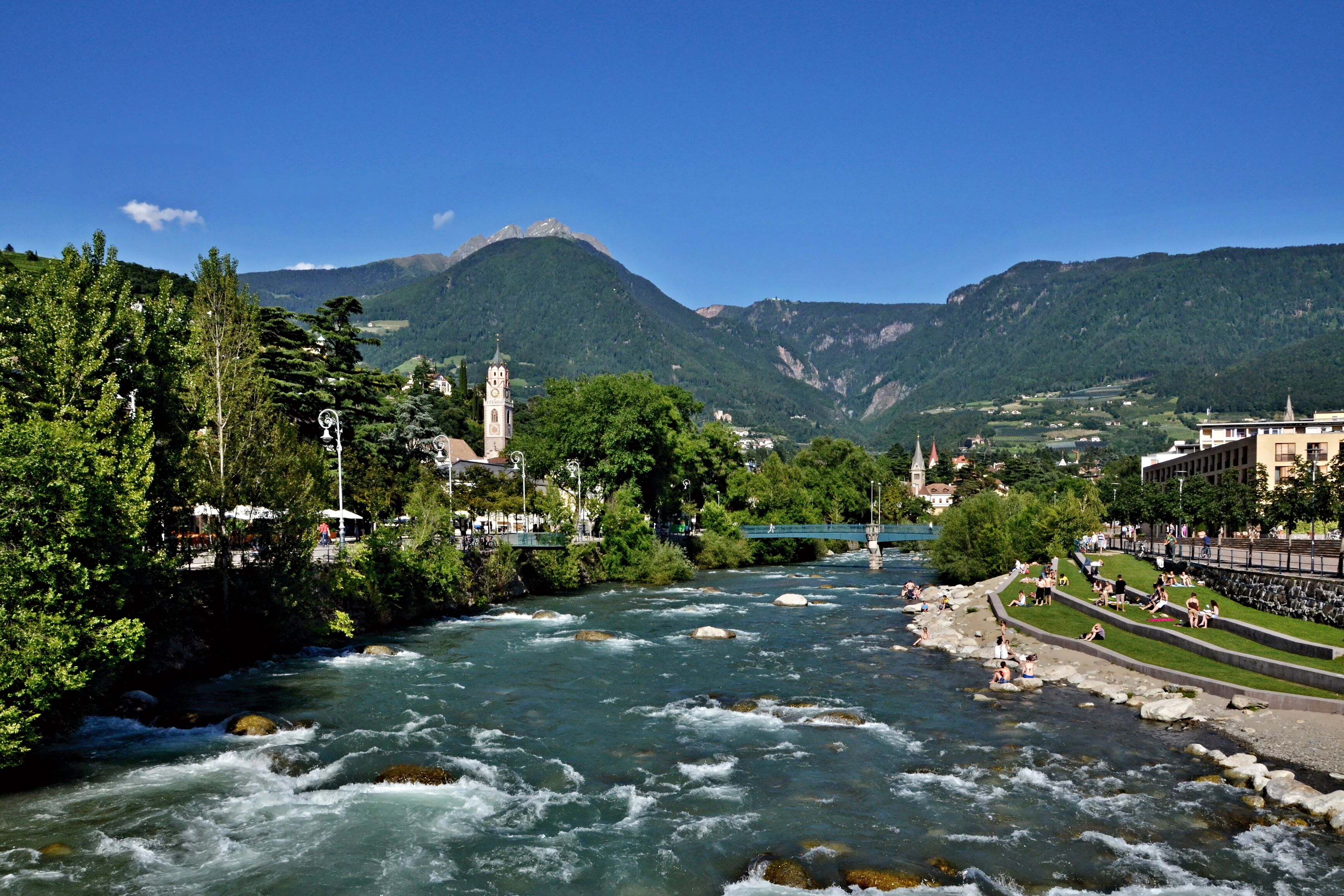 A river flowing through a valley with mountains in the background, a bridge, and a park with people.