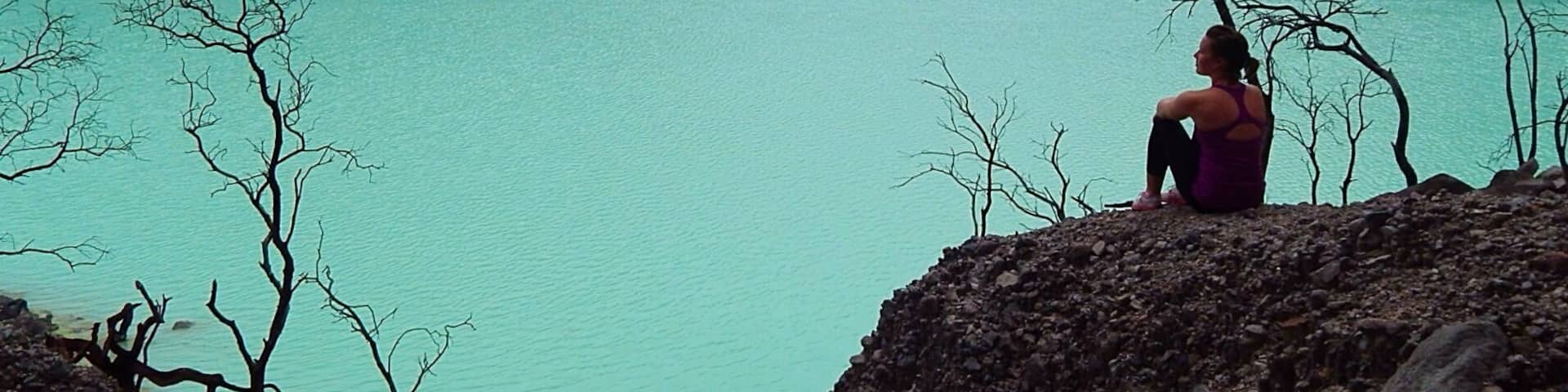 A person sitting on a rocky outcrop overlooking a turquoise lake with mountains in the background.