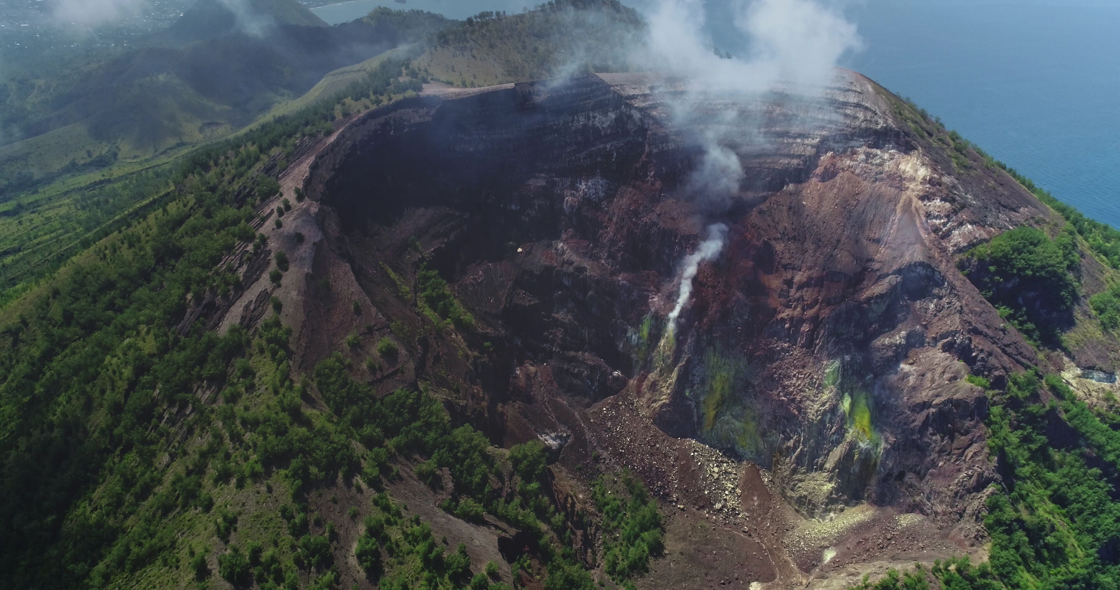 White smoke is rising from the crater of Mount Iya volcano, located near Ende on Flores island, Indonesia, surrounded by lush green vegetation and overlooking the ocean