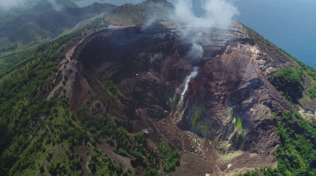 White smoke is rising from the crater of Mount Iya volcano, located near Ende on Flores island, Indonesia, surrounded by lush green vegetation and overlooking the ocean