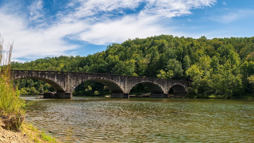 A stone bridge over a river with a forested hillside in the background.