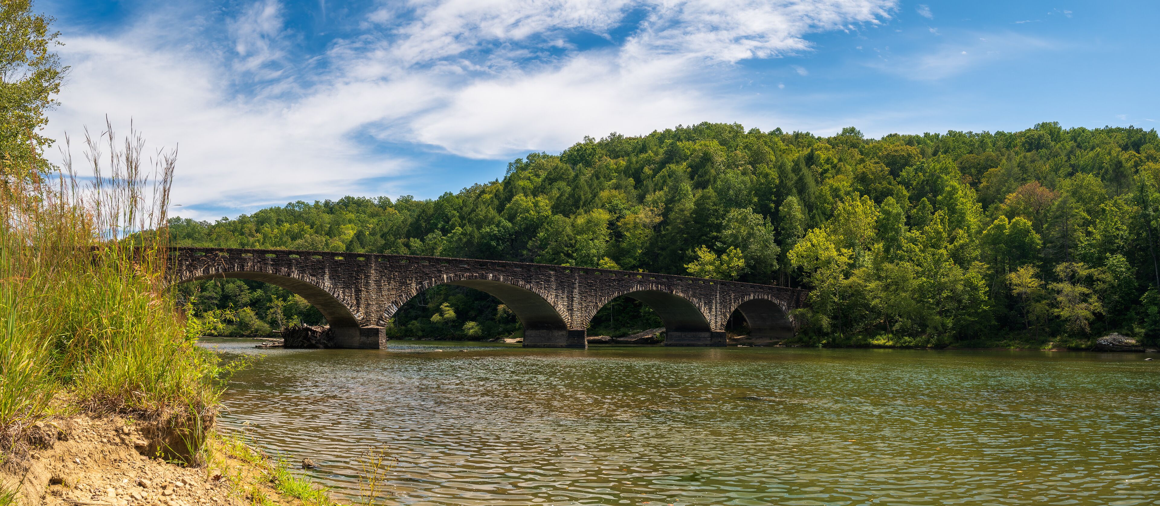 A stone bridge over a river with a forested hillside in the background.