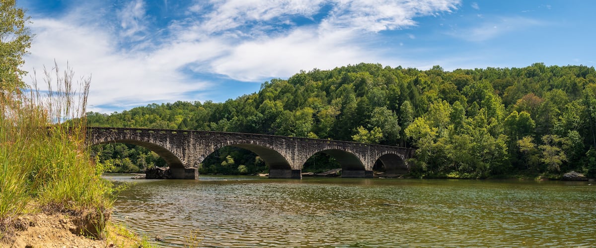 A stone bridge over a river with a forested hillside in the background.