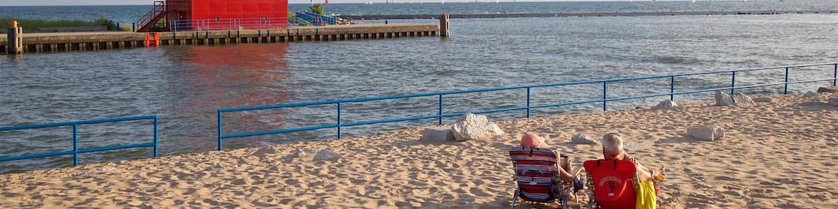Ein roter Leuchtturm auf einem Steg, ein Sandstrand und zwei Personen, die auf Strandstühlen sitzen.