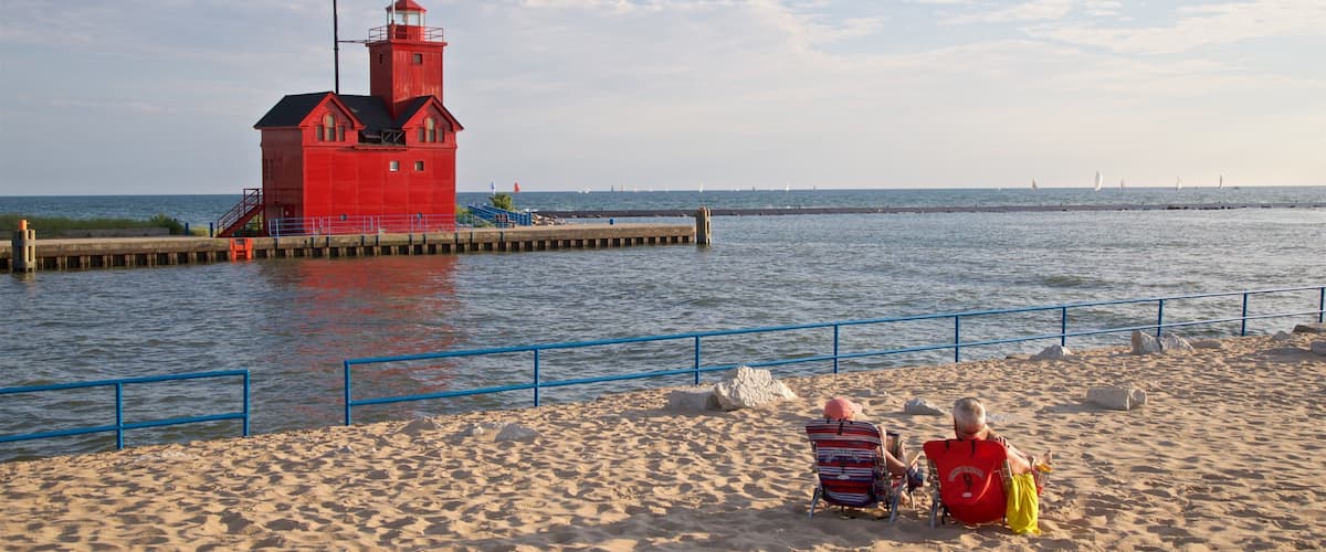 Un faro rojo sobre un muelle, una playa de arena y dos personas sentadas en sillas de playa.