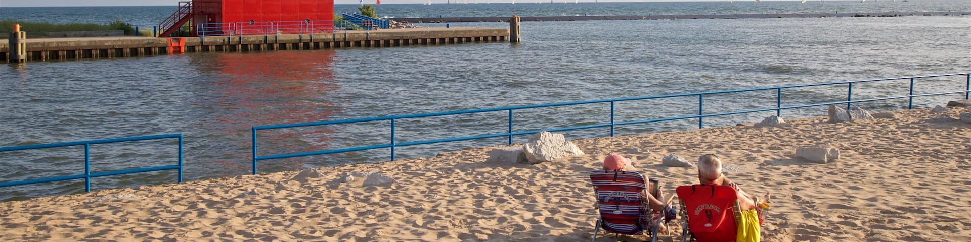 A red lighthouse on a pier, a sandy beach, and two people sitting on beach chairs.