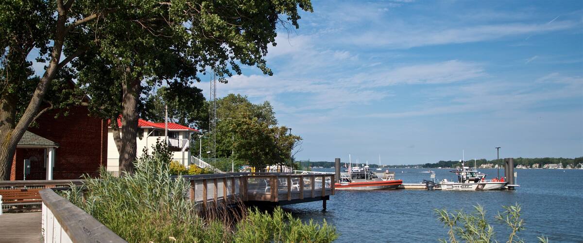 A wooden dock extending into a calm body of water, with boats docked nearby and a building with a red roof in the background.