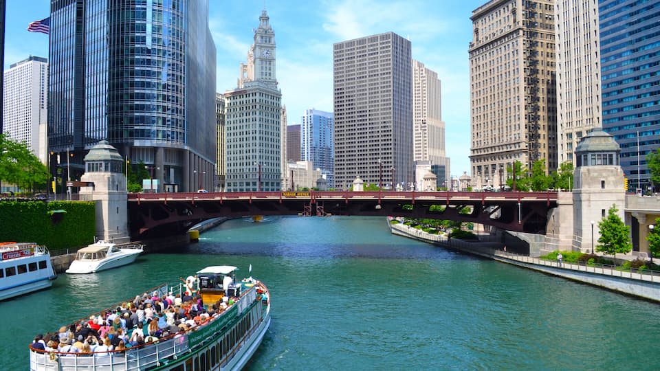 A river with a boat carrying passengers, surrounded by tall buildings and a bridge.