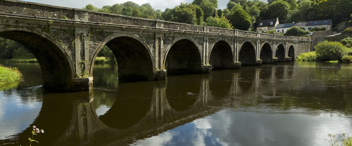 A stone bridge with multiple arches spanning a river, surrounded by greenery and houses in the background.