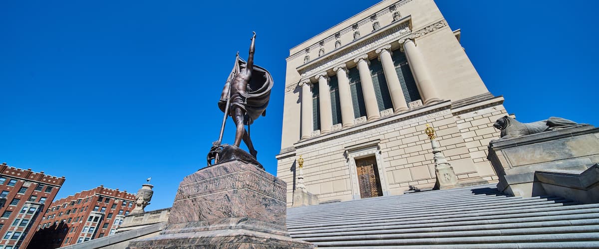 A statue on a pedestal in front of a building with columns and a clock tower.