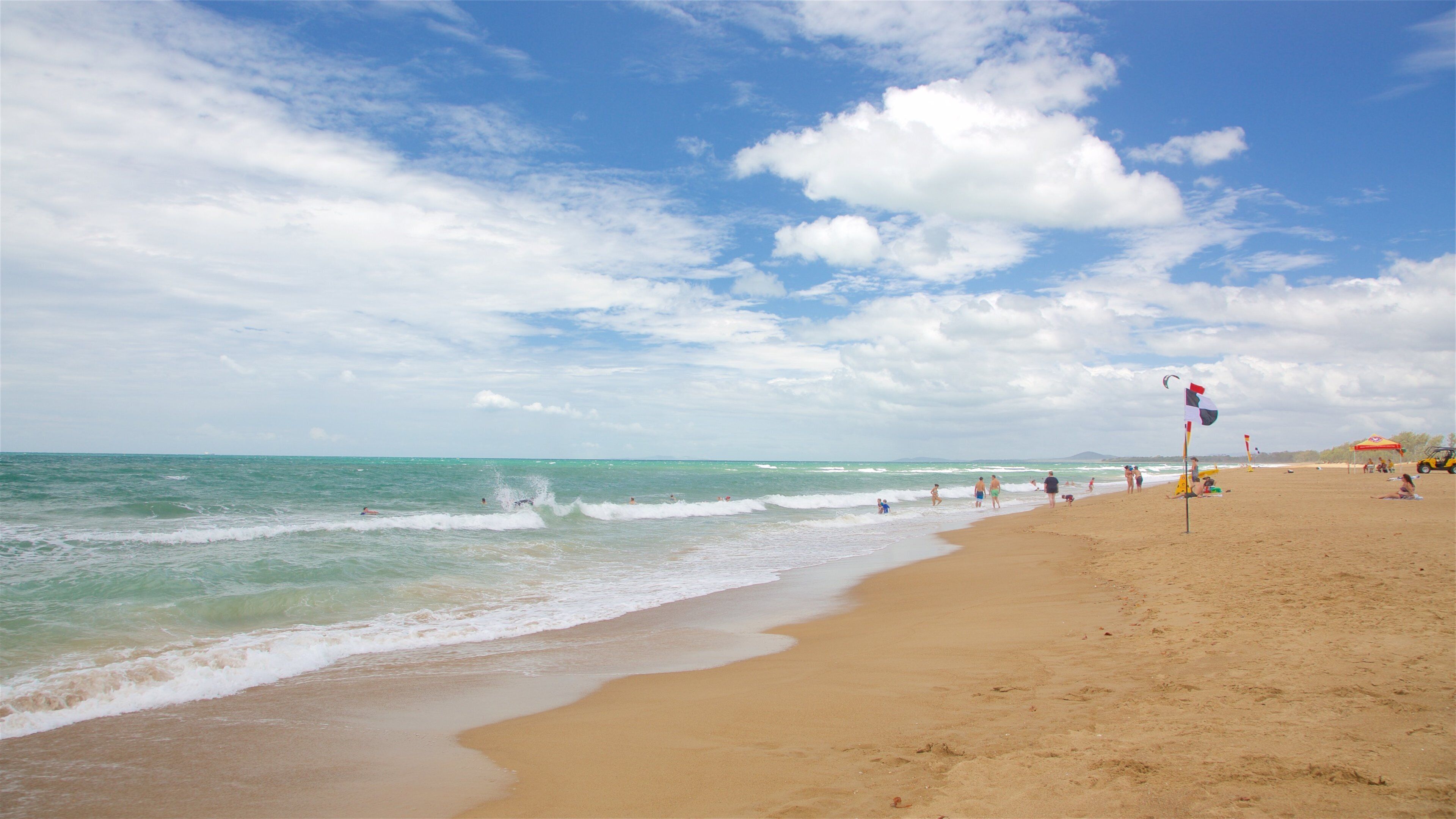 A sandy beach with clear turquoise water, a few people swimming, and a lifeguard flag.