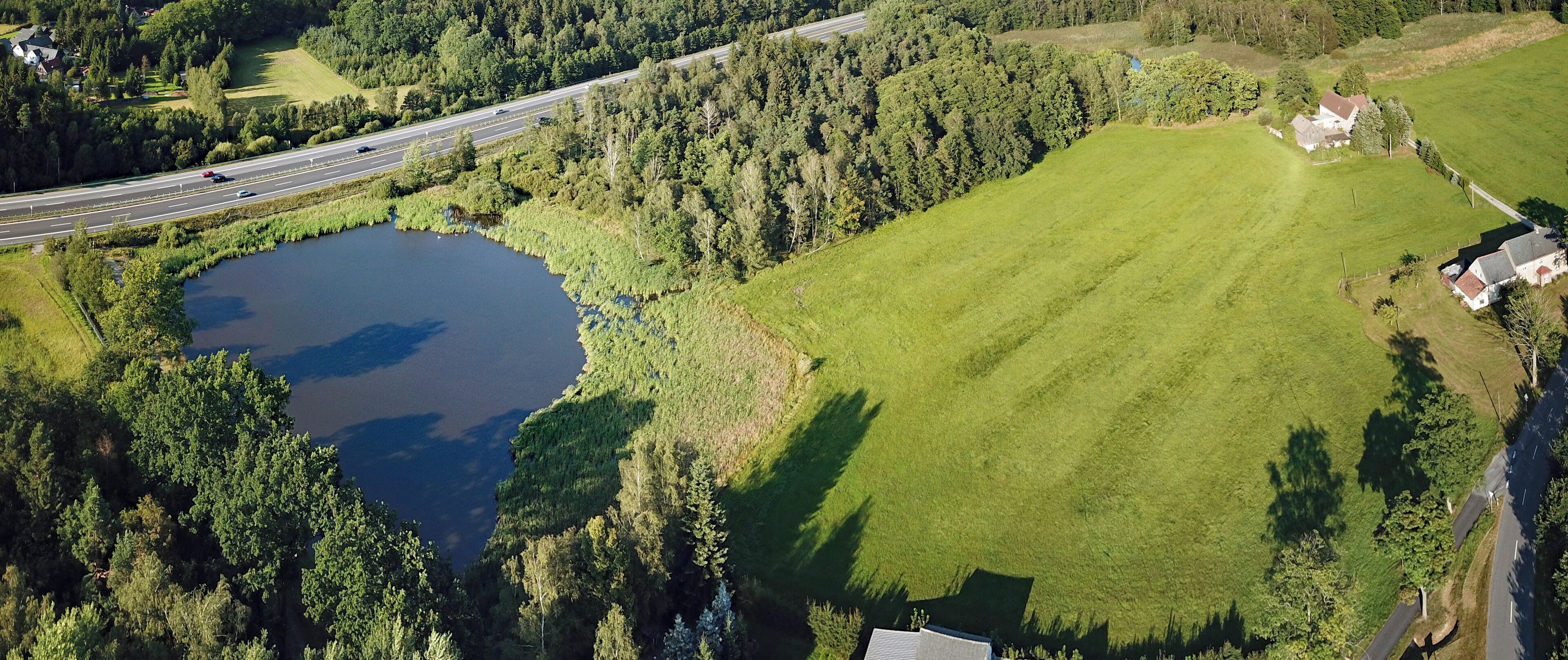 An aerial view of a lake surrounded by dense forest, a highway, and a large grassy field.