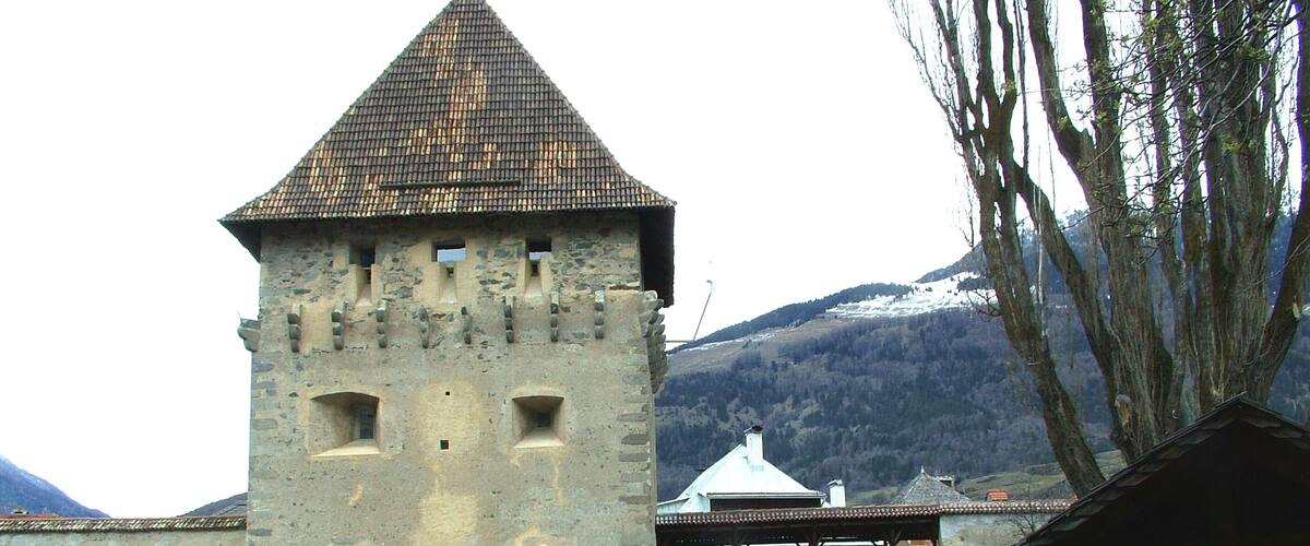 A historic stone tower with a tiled roof, a bridge, and a mountainous backdrop.