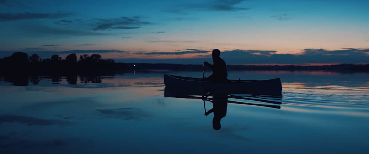 A silhouette of a person in a canoe on a calm lake at dusk with a sunset in the background.