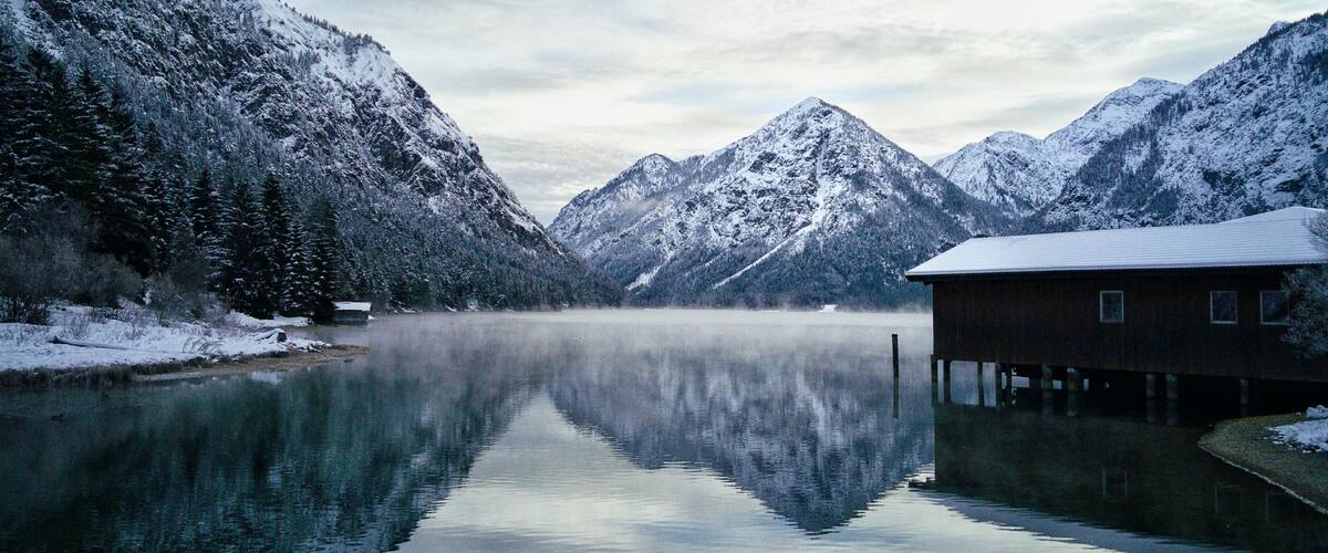 A snow-covered mountain range, a calm lake, and a wooden cabin.