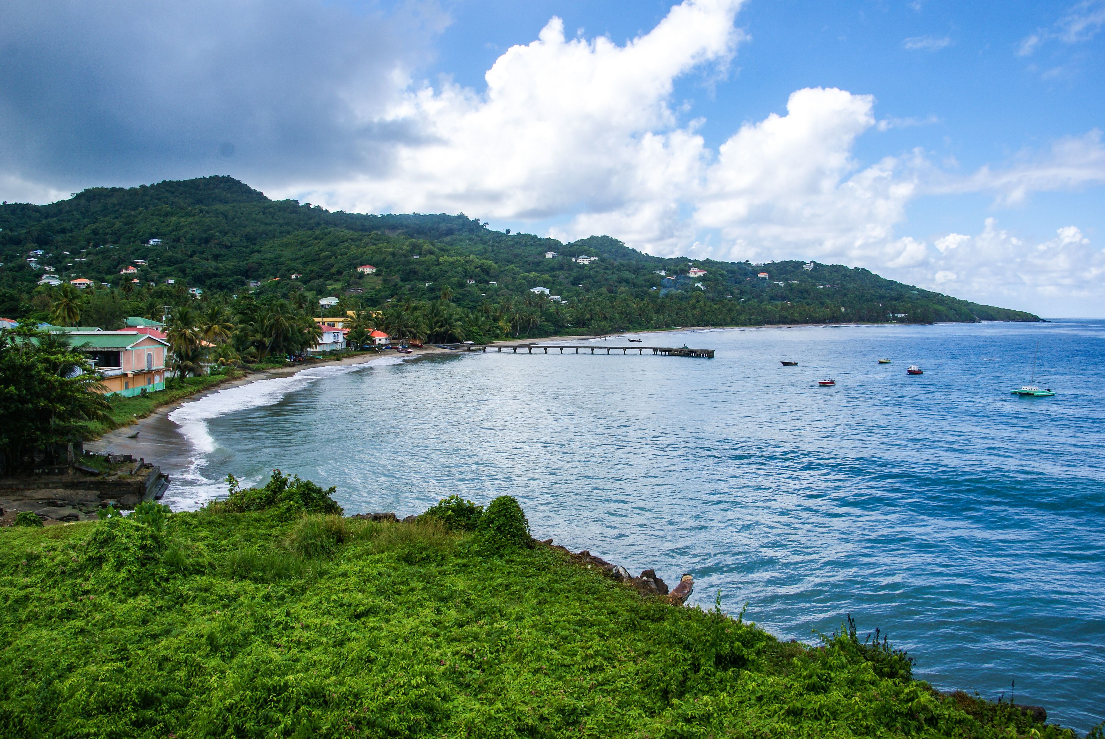 A coastal landscape with a pier extending into the sea, surrounded by lush greenery and a backdrop of hills.