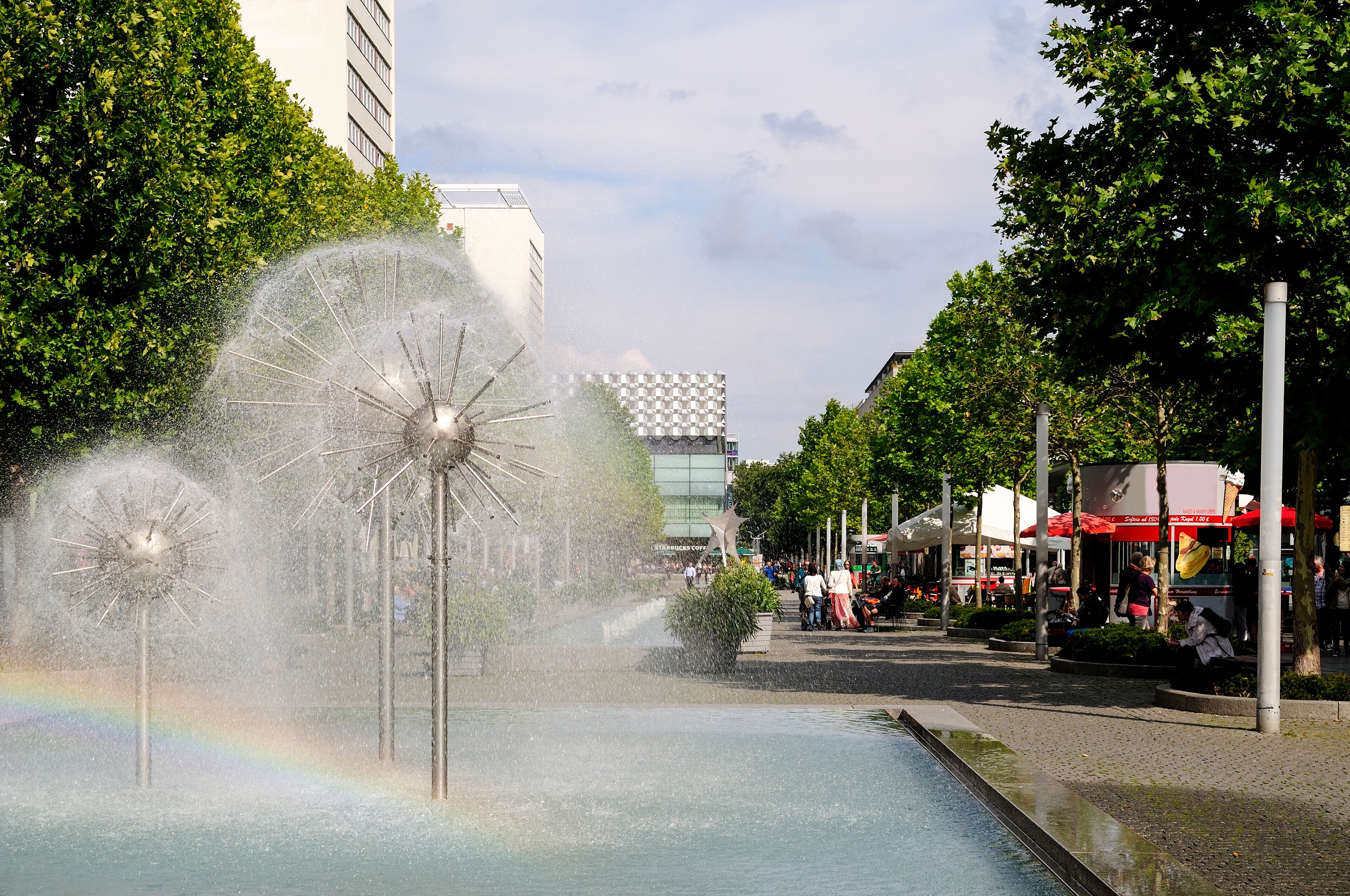 A fountain with water jets, a rainbow, and a checkered building in the background.