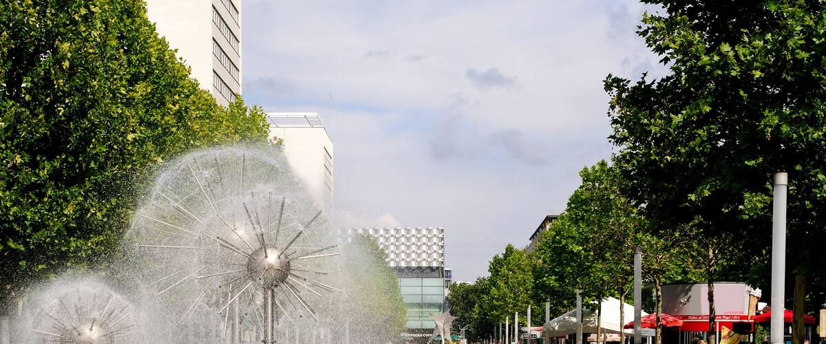 A fountain with water jets, a rainbow, and a checkered building in the background.