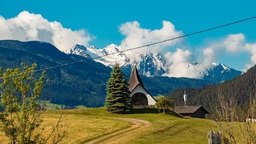 Een kerk met een kruis bovenop, omgeven door een berglandschap met besneeuwde toppen en een helderblauwe lucht.