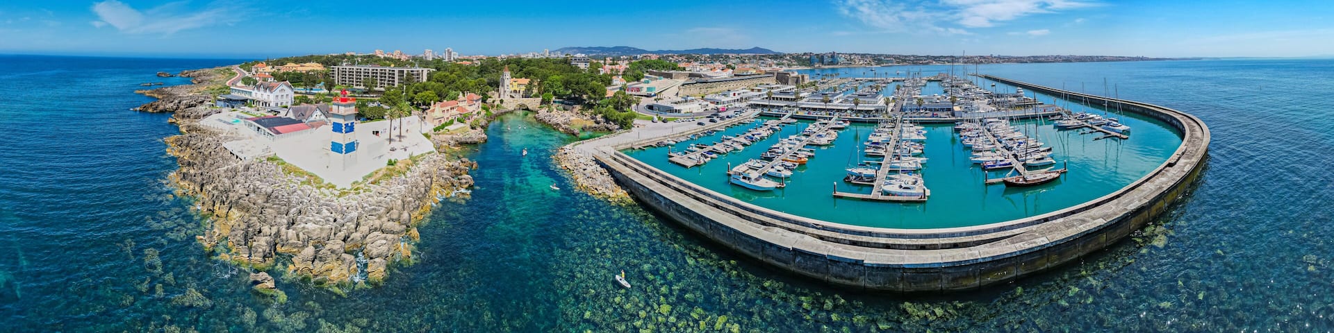 A coastal town with a marina, clear blue waters, rocky shoreline, and a lighthouse.