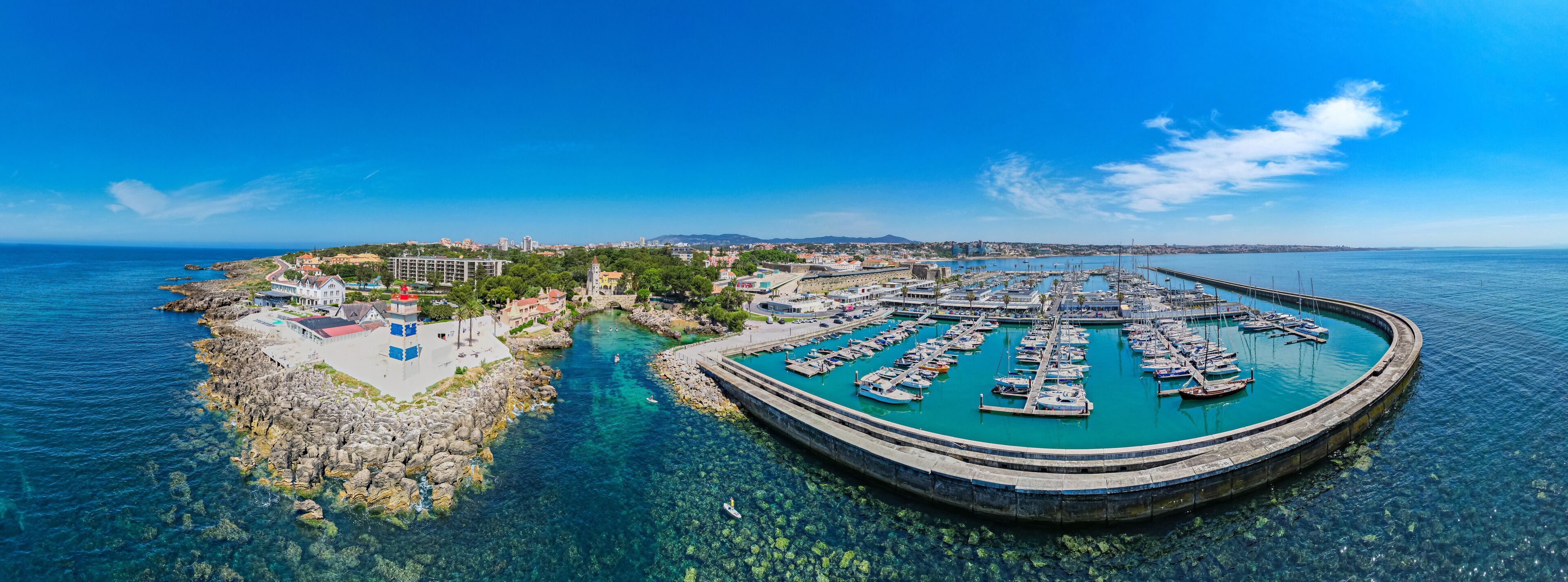 A coastal town with a marina, clear blue waters, rocky shoreline, and a lighthouse.