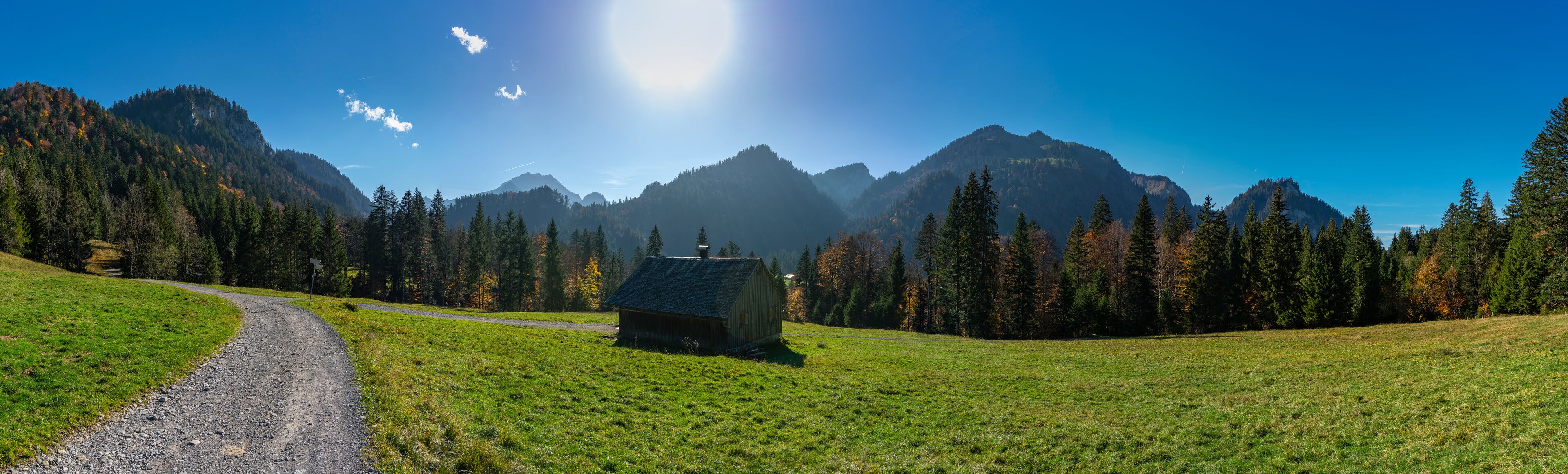 A rural landscape with a dirt road, a small wooden cabin, and a mountain range under a clear blue sky.